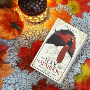 The Fool and the Sparrow book cover mockup, surrounded by fall leaves, a velvet pumpkin and a shining acorn lantern