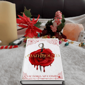 A mockup of Oathbound, with a vivid red rose with a sword through it, on a background of a fuzzy white blanket, a candy cane, a pillar candle, a sparkly fake poinsettia, a snowman, a Himalayan salt votive, some stones and mini Christmas lights